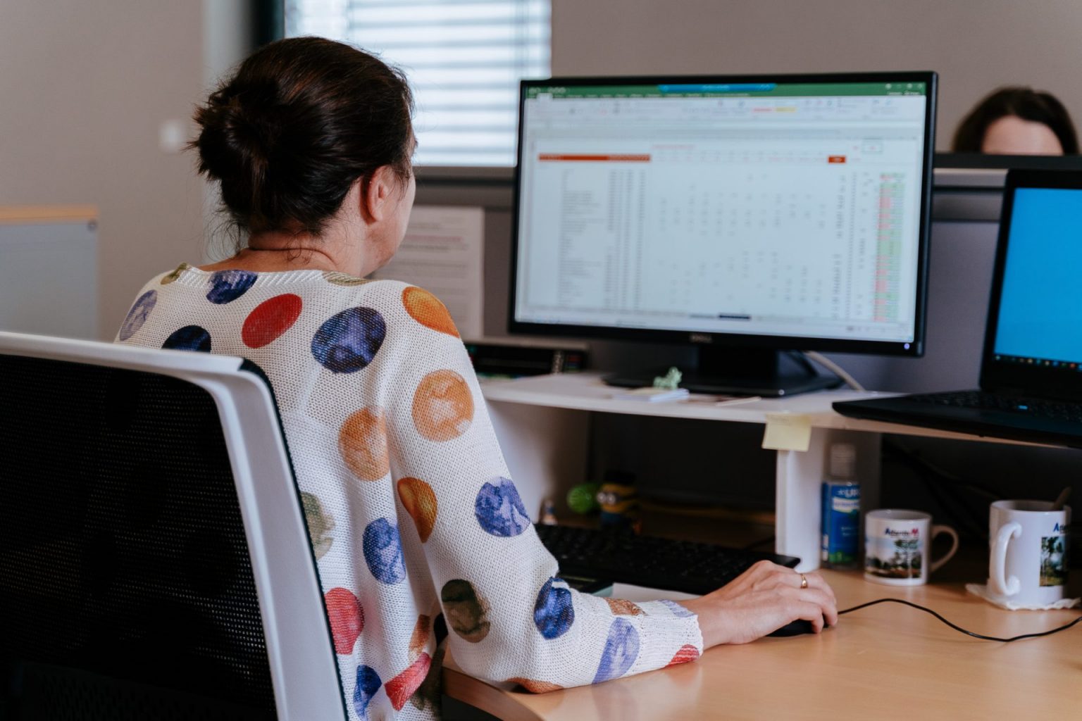 Photographie d'une femme de dos portant un pull coloré, devant son écran d'ordinateur à son poste de travail